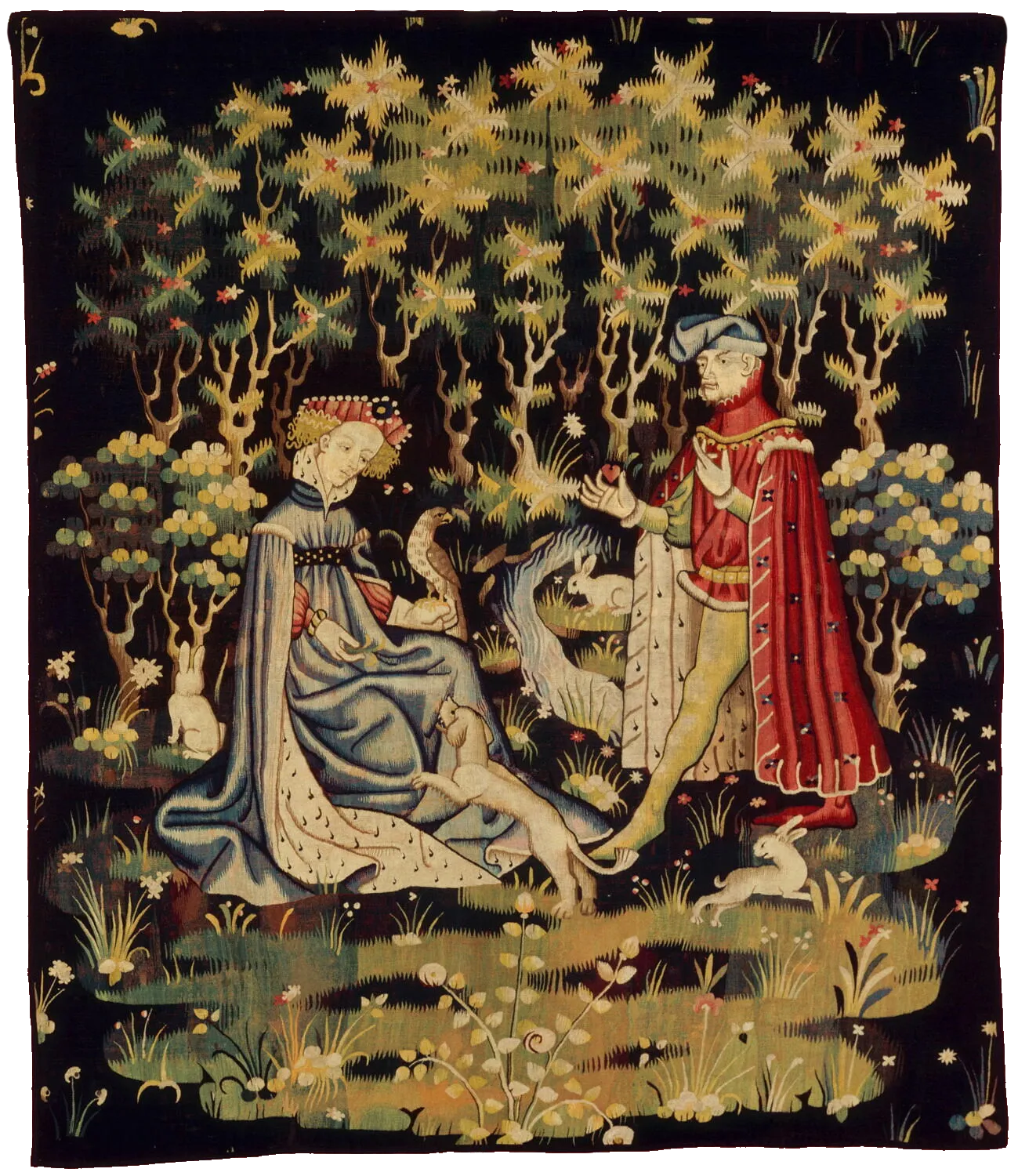 L'image est une tapisserie représentant un homme et une femme dans un contexte champêtre. La femme est agenouillée, un faucon est posé sur sa main gauche alors que de la gauche elle joue avec un petit chien. L'homme s'avance vers elle avec un petit objet en forme de petit coeur dans sa main droite.