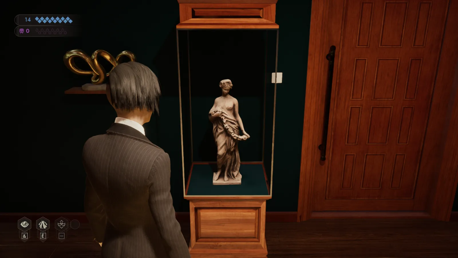 L'image est une capture d'écran du jeu vidéo Vampire: The Masquerade – Swansong. On y voit le personnage joueur de dos, debout dans une pièce aux murs couleur vert sapin. Face au personnage joueur, contre le mur, une vitrine jouxte une porte en bois claire. La vitrine renferme une statuette représentant une jeune femme.