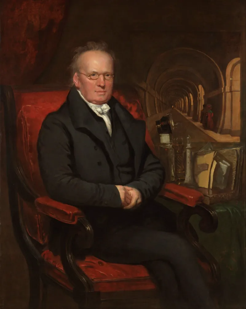 Portrait de Sir Marc Isambard Brunel. Un homme d'une soixantaine d'années en costume et portant des lunettes est assis dans un fauteuil tapissé de tissu rouge. Il regarde directement le spectateur, il a les jambes croisées et les mains jointes au niveau du ventre. A côté de lui se trouve une table recouverte de tissu vert et de divers objets. A l'arrière plan la perspective s'enfonce dans un long tunnel piéton.