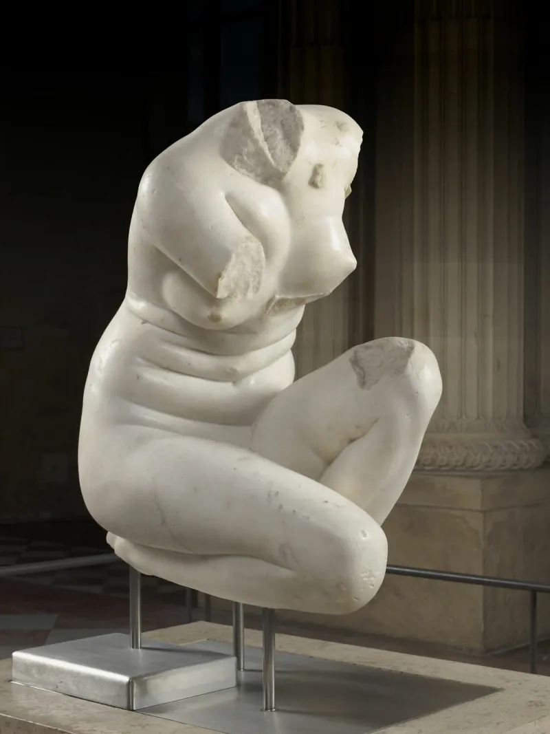 Photographie d'une sculpture antique représentant une femme nue accroupie. La sculpture est très endommagée : il lui manque la tête et les deux bras.