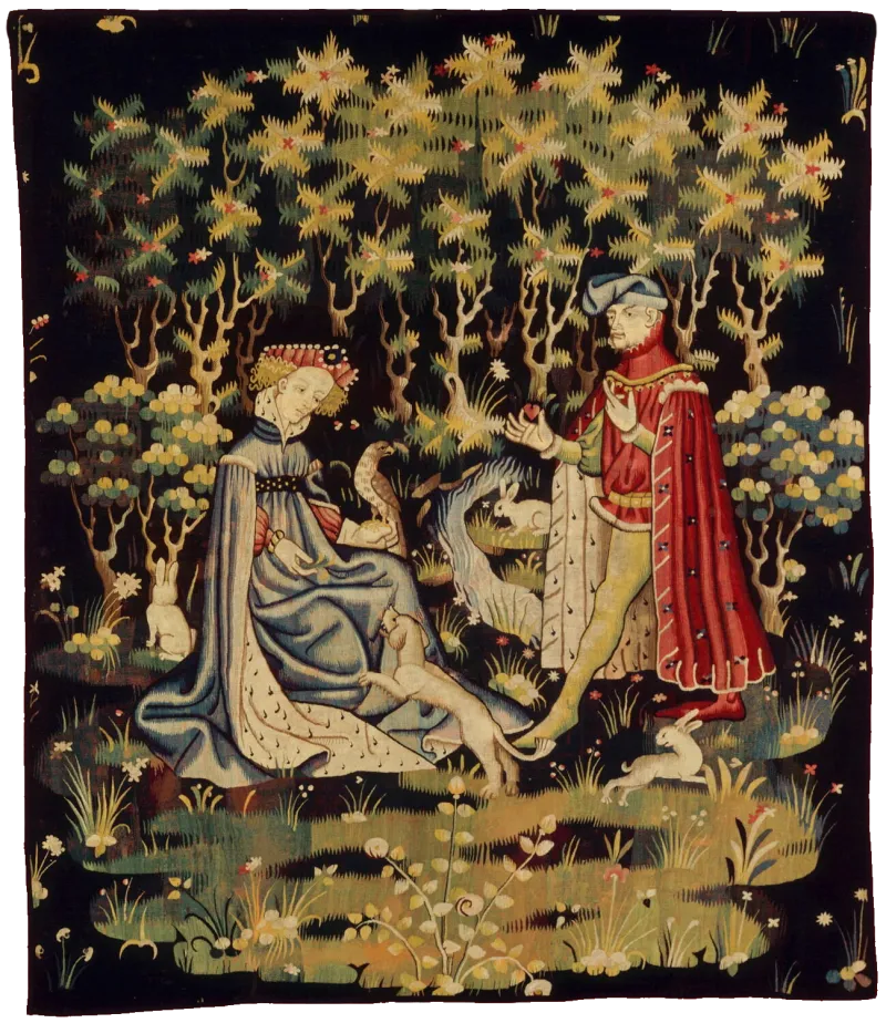 L'image est une tapisserie représentant un homme et une femme dans un contexte champêtre. La femme est agenouillée, un faucon est posé sur sa main gauche alors que de la gauche elle joue avec un petit chien. L'homme s'avance vers elle avec un petit objet en forme de petit coeur dans sa main droite.