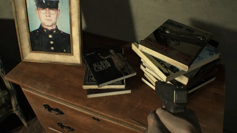 L'image est une capture d'écran du jeu Resident Evil 7. La caméra, qui adopte le point de vue du personnage joueur, nous montre une table sur laquelle est posée une photographie dans son cadre, ainsi que deux piles de livres. Au centre de la table et de la capture d'écran, la couverture de l'ouvrage en haut de la plus petite des piles présente une image d'une statue antique.
