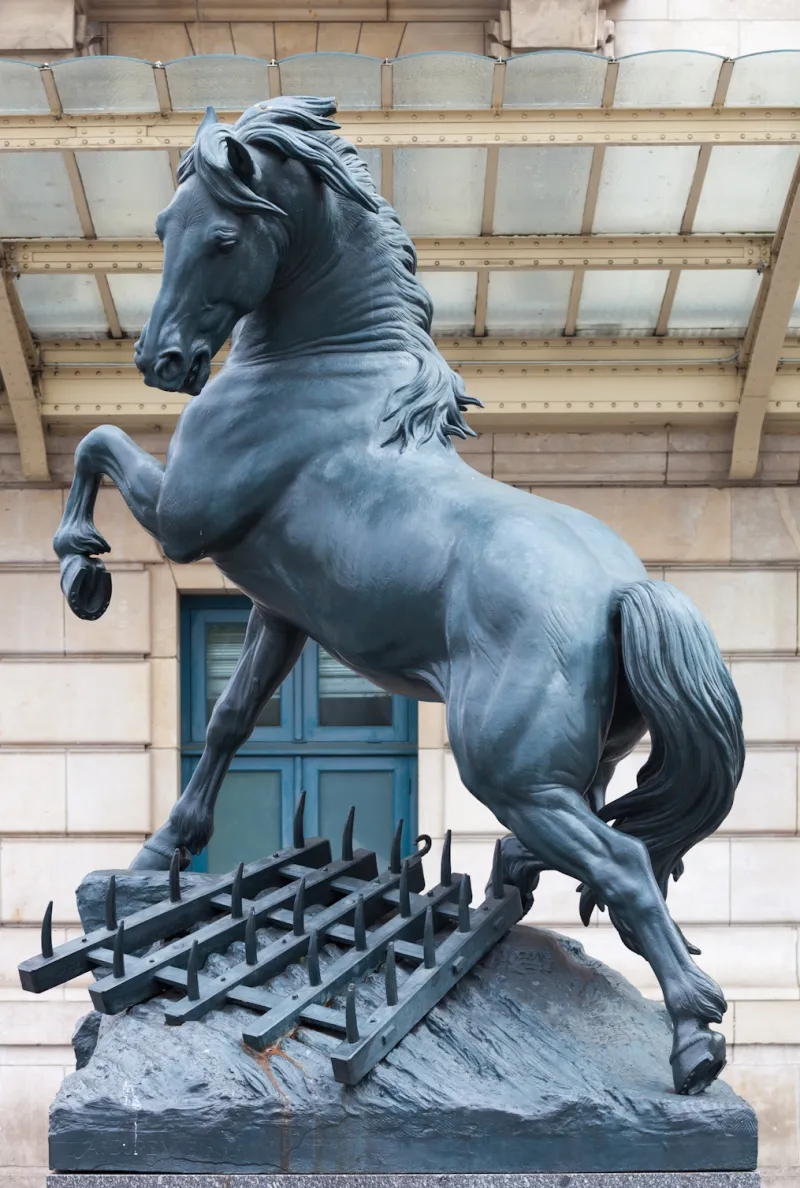 L'image est une photographie d'une grande sculpture en bronze représentant un cheval enjambant une herse.