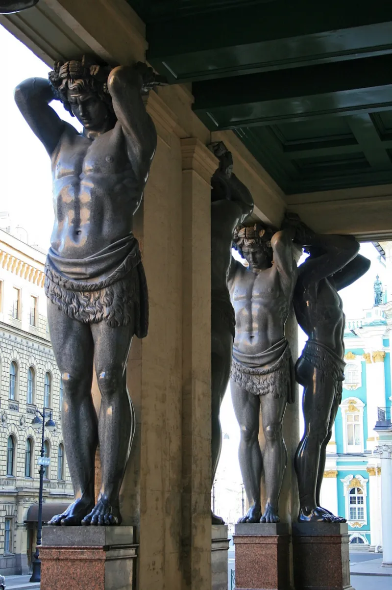 L'image est une photographie prise depuis l'intérieur d'un portique dont les piliers sont ornés de grands atlantes sculptés dans une pierre sombre.