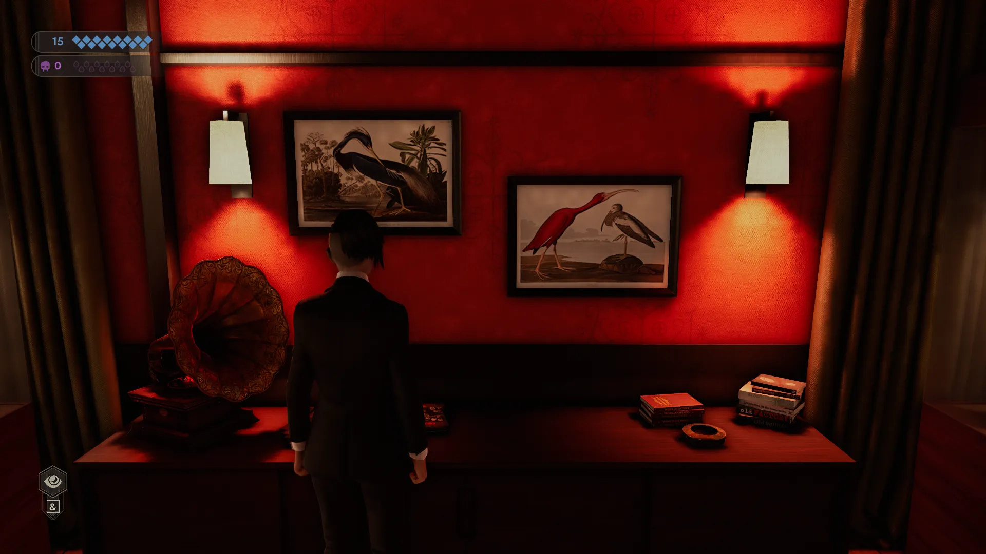 L'image est une capture d'écran du jeu vidéo Vampire: The Masquerade – Swansong. On y voit le personnage joueur de dos, face à un mur tapissé de papier peint rouge. Plusieurs objets sont posés devant lui sur un large meuble : un gramophone, un cendrier et des piles de livres. Au mur, entre deux appliques, sont accrochés deux estampes colorées représentant des oiseaux.