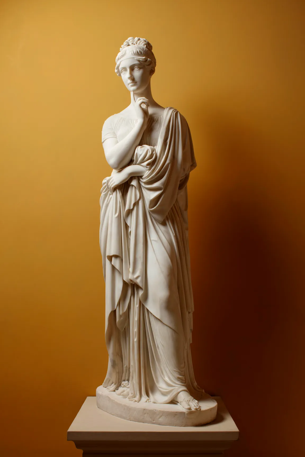 L'image est une photo d'une sculpture en marbre disposée devant un mur peint d'une couleur orangée. La sculpture représente une jeune femme debout, vêtue à l'antique, le bras gauche ramené contre son ventre, et la main droite près de son cou, son index et son majeur touchant son menton dans une attitude de réflexion.