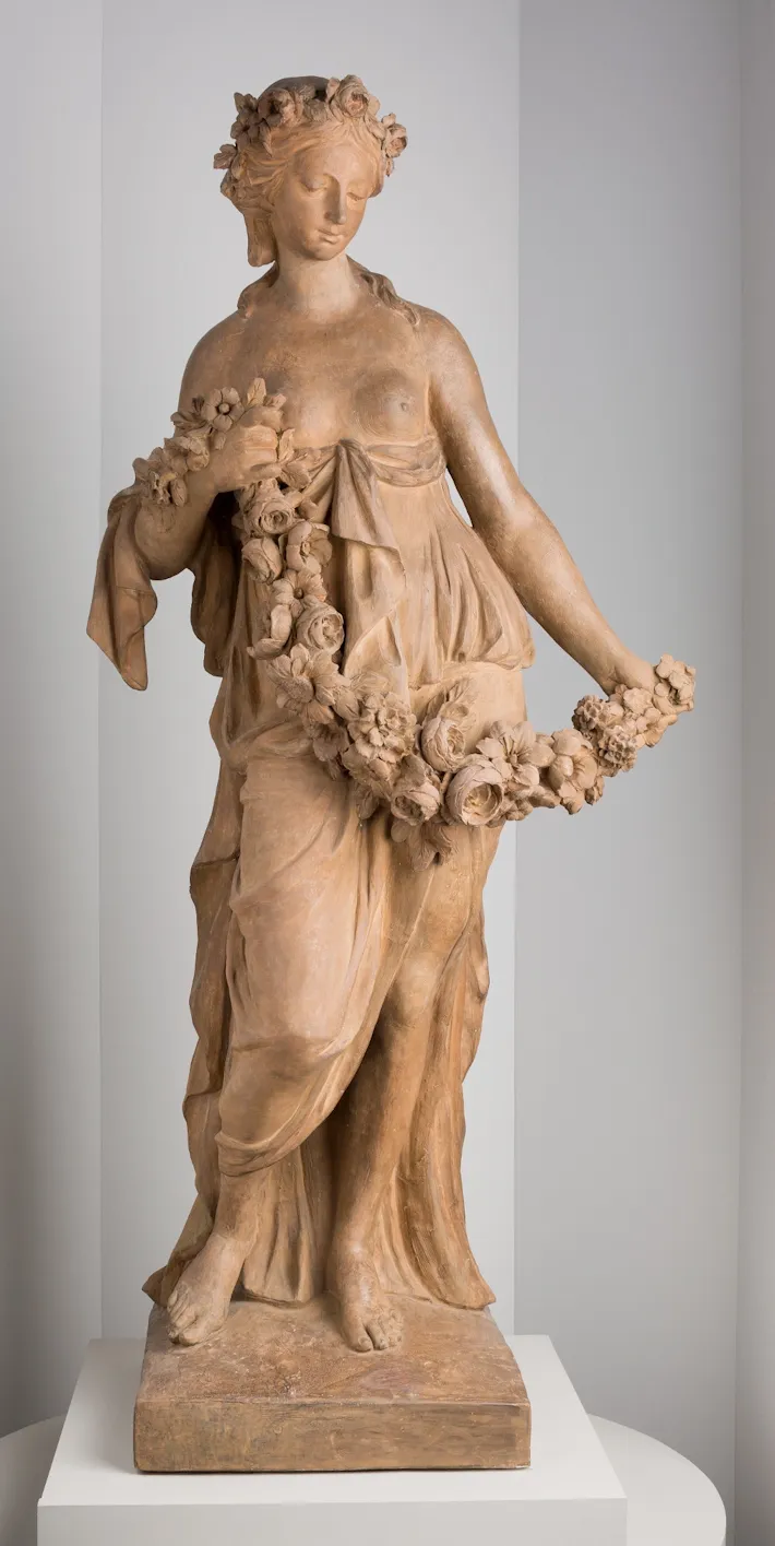 L'image est une photographie d'une sculpture en terre cuite. Elle représente une jeune femme debout, vêtue à l'antique, la poitrine dénudée, tenant dans ses mains une guirlande de fleurs. Elle est également coiffée d'une couronne de fleurs.