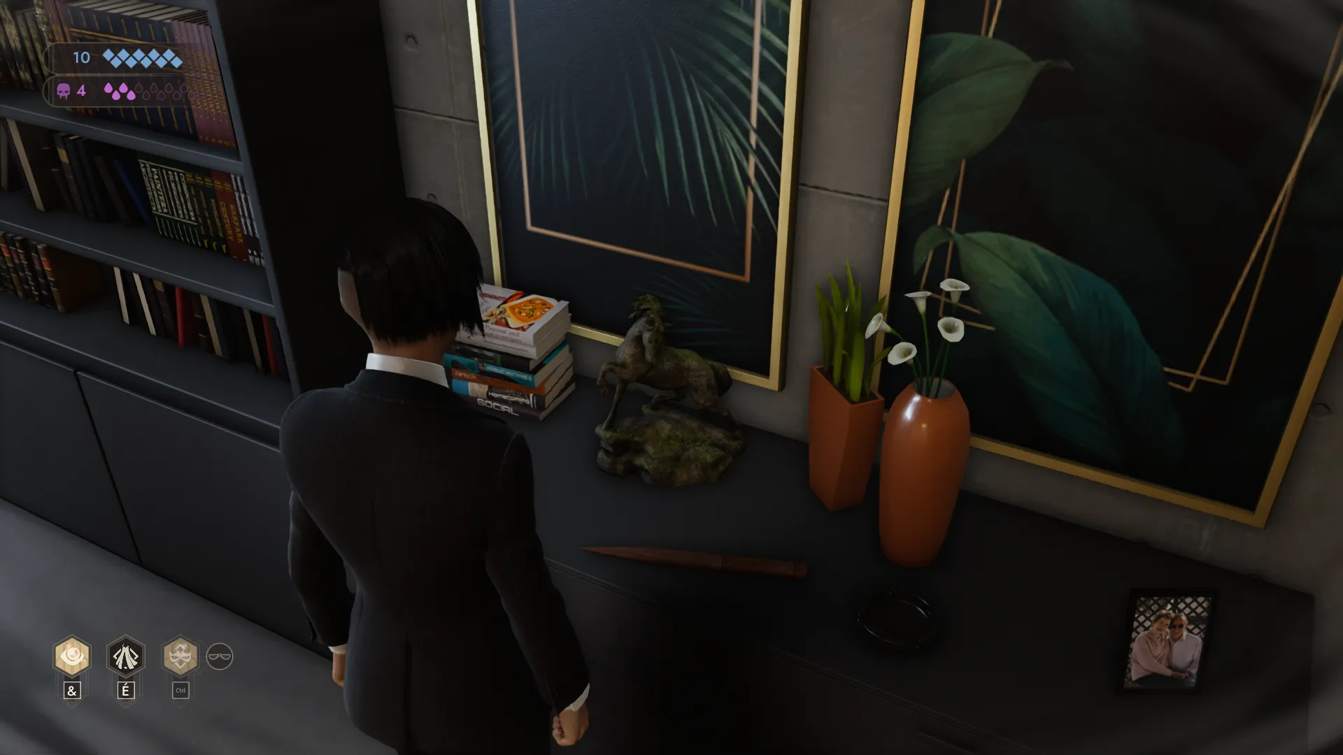 L'image est une capture d'écran du jeu vidéo Vampire: The Masquerade – Swansong. On y voit le personnage joueur de dos, debout face à un meuble bas adossé à un mur. Divers objets sont disposés sur le meuble : deux plantes en pot, une photographie encadrée, une pile de livres, un poignard en bois ainsi qu'une petite sculpture en bronze représentant un cheval.